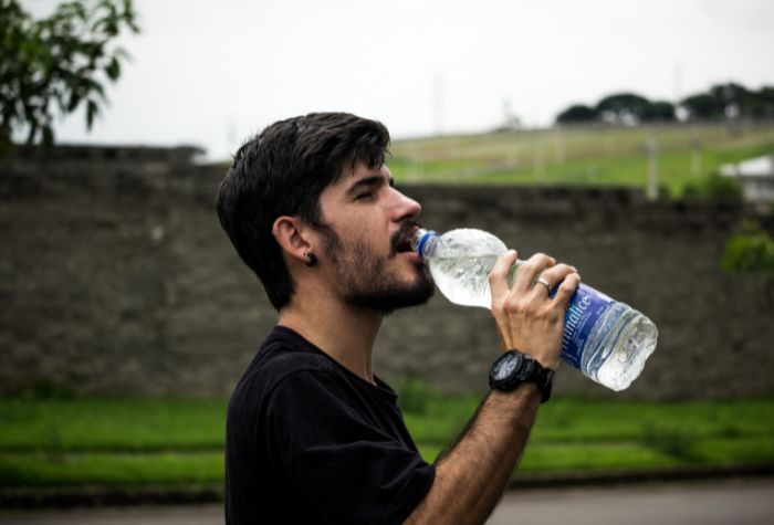 A dark-haired young man drinking a large bottled water in front of a grassy background